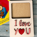 A wooden photo frame with the text 'I love you' arranged in a playful font using cut-out letters, with a red heart replacing the word 'you'.