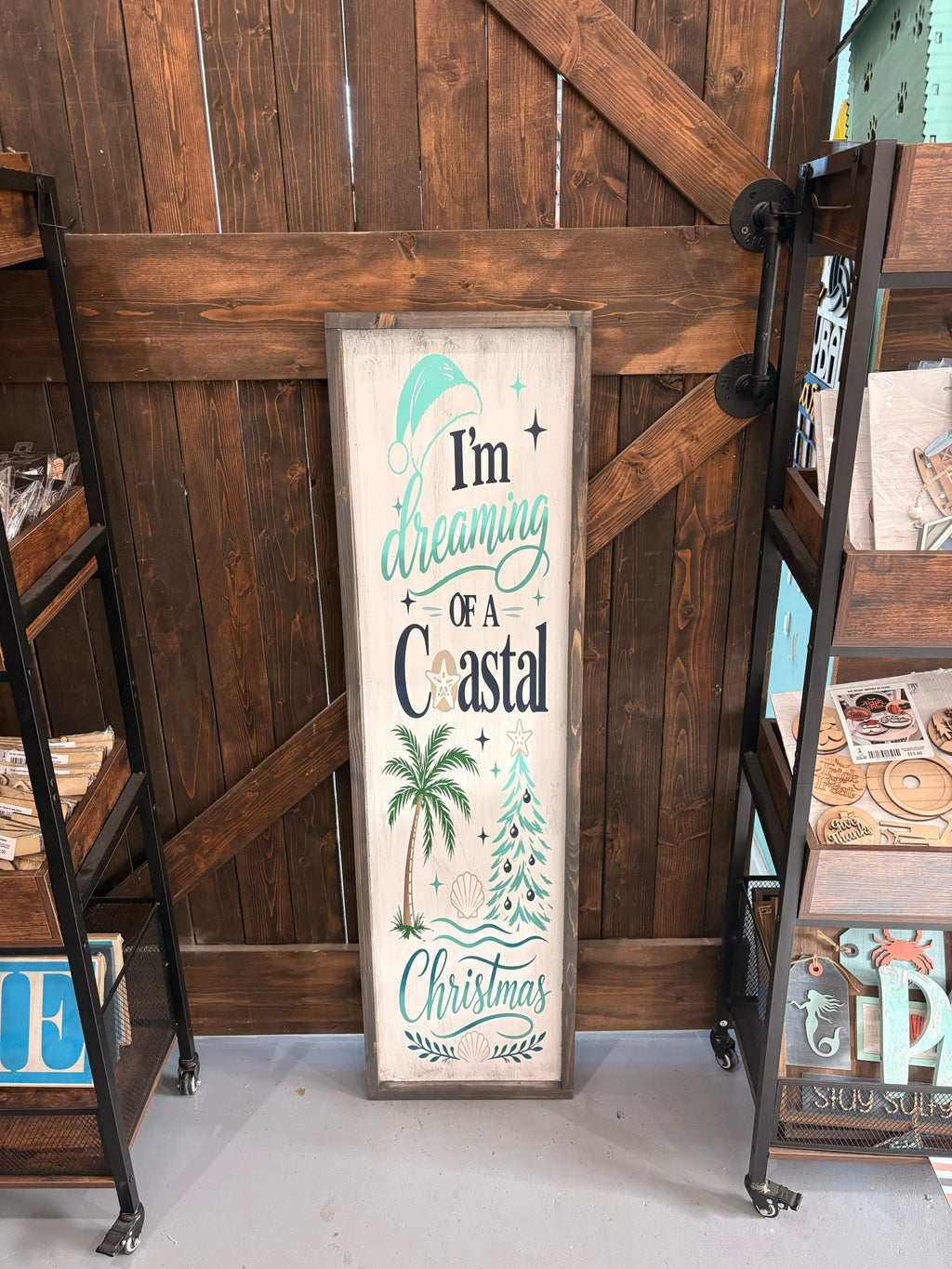 A vertical rectangular wooden sign with a distressed cream background and rustic brown frame, featuring the phrase “I’m dreaming of a coastal Christmas” in teal and dark lettering, with illustrated palm trees and a Christmas tree. The sign is leaning against a dark wood barn-style door between shelving units.