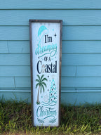 A vertical rectangular wooden sign with a rustic dark brown frame, featuring the text 'I'm dreaming of a Coastal Christmas' in teal and black lettering, decorated with illustrations of a palm tree, Christmas tree, and seashell, leaning against a light blue wooden wall on grass.