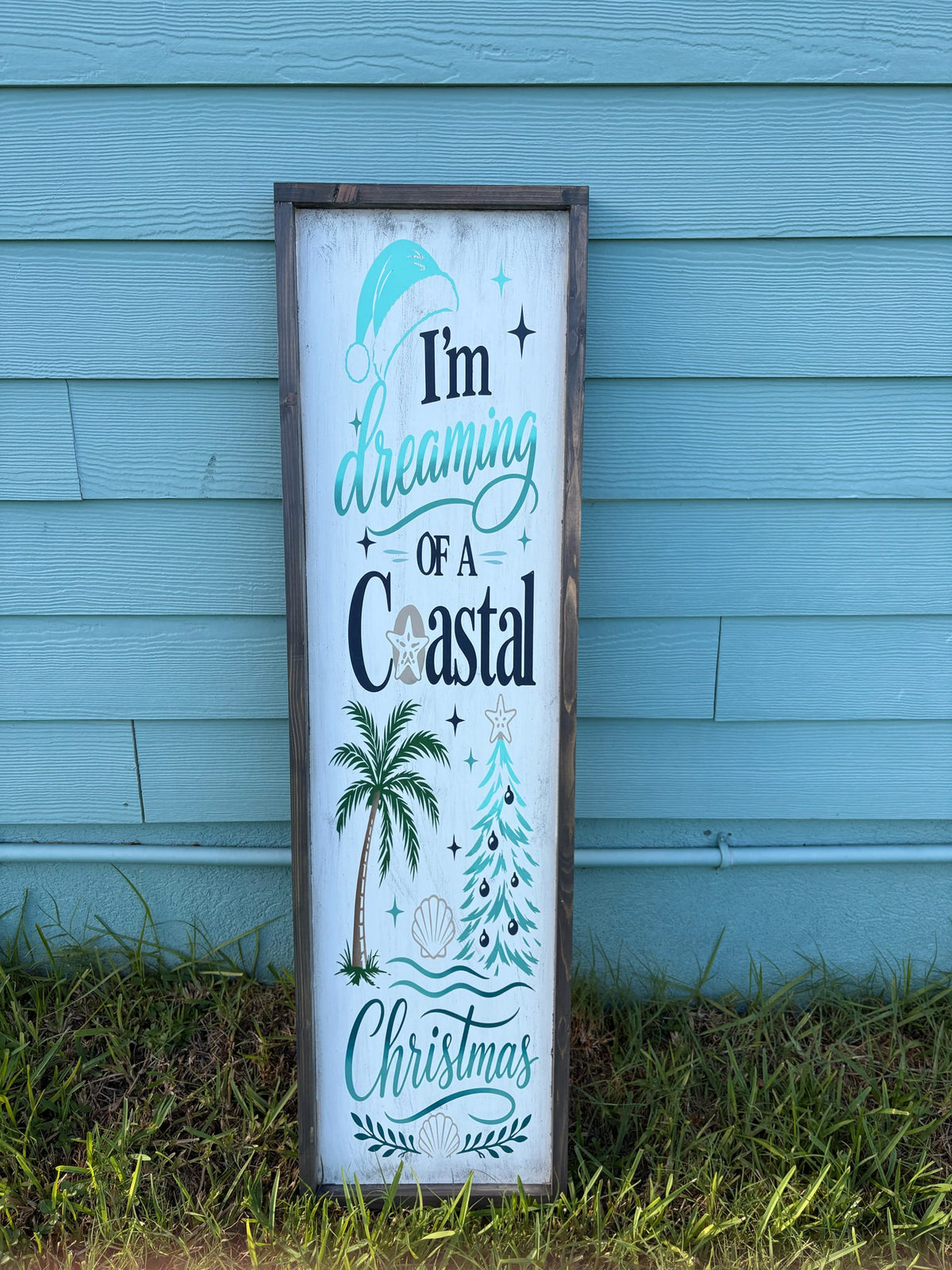 A vertical rectangular wooden sign with a rustic dark brown frame, featuring the text 'I'm dreaming of a Coastal Christmas' in teal and black lettering, decorated with illustrations of a palm tree, Christmas tree, and seashell, leaning against a light blue wooden wall on grass.