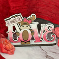 A shelf sitter decoration featuring a moose with the word 'love' formed from its body, in shades of pink and brown, with a 'It Moose Be Love' text above it.