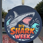 Decorative sign with shark illustration and 'It's Shark Week' text on a glass door.