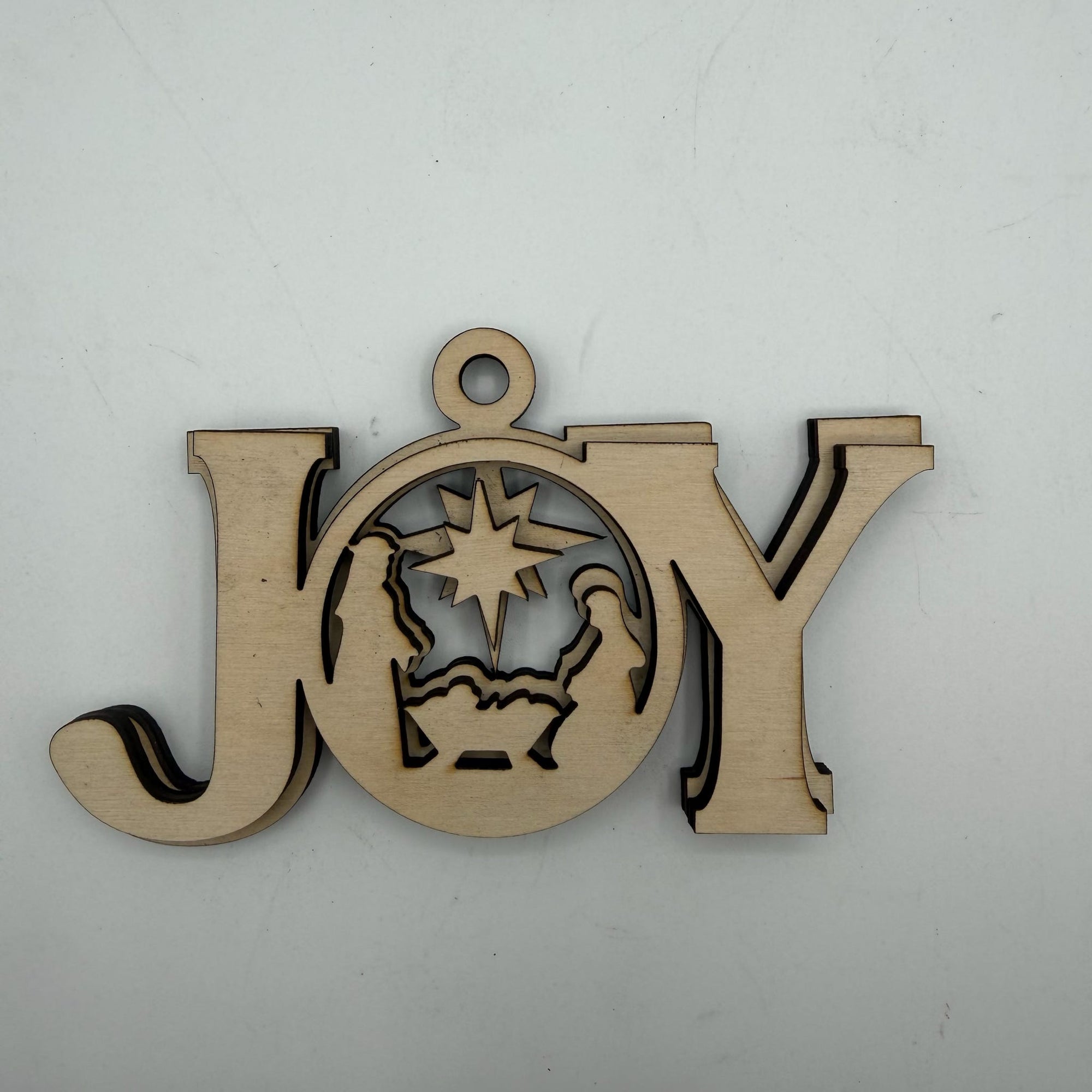 A laser-cut wooden Christmas ornament spelling out 'JOY' with a nativity scene cutout in the center of the letter O, shown on a plain light gray background.