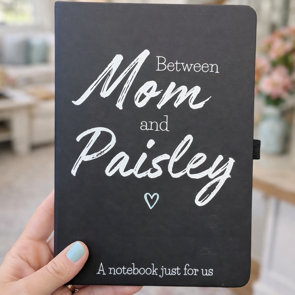 Black notebook with 'Between Mom and Paisley' text held in a hand, blurred indoor background