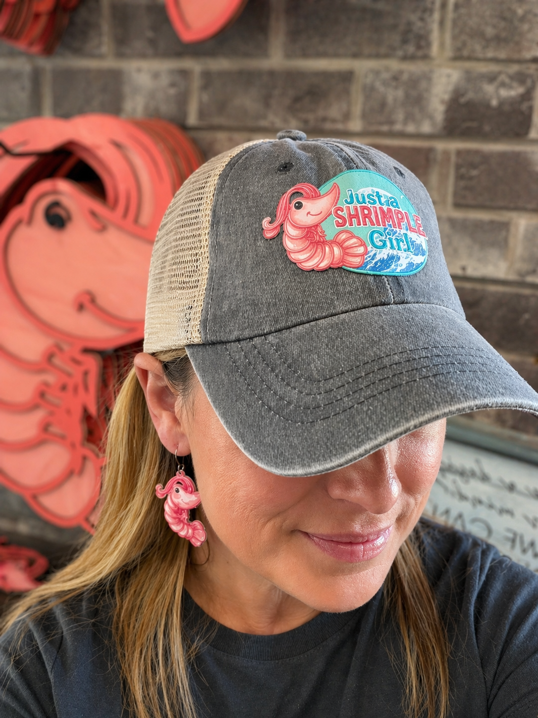 Person wearing a cap with a 'Just a Shrimp Girl' logo against a brick wall.