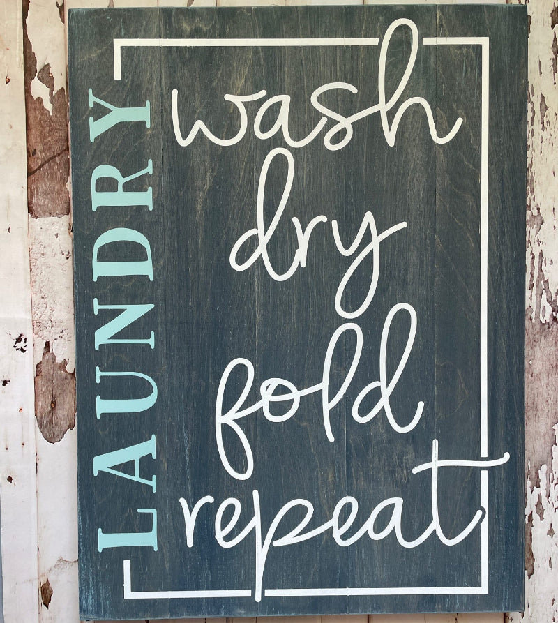 A laundry room sign with the words 'wash', 'dry', 'fold', and 'repeat' arranged in a rectangular frame against a wooden background.