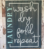 A laundry room sign with the words 'wash', 'dry', 'fold', and 'repeat' arranged in a rectangular frame against a wooden background.