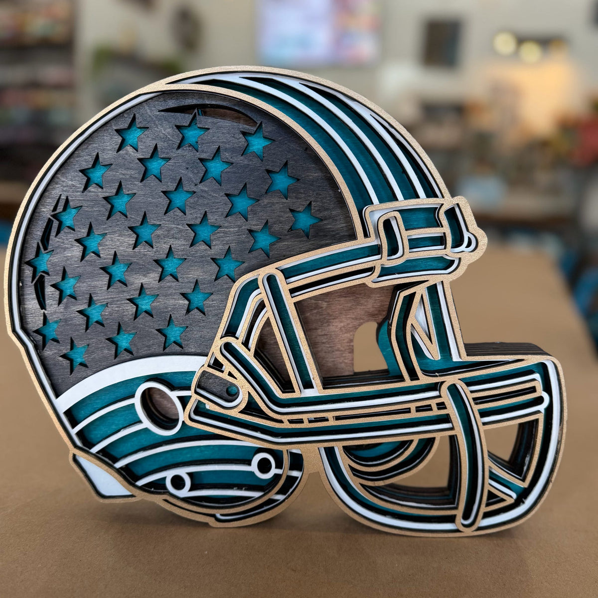 A wooden American football helmet cutout with a distressed dark gray wood finish, teal blue inlay, and white stripes, set against a blurred indoor background.