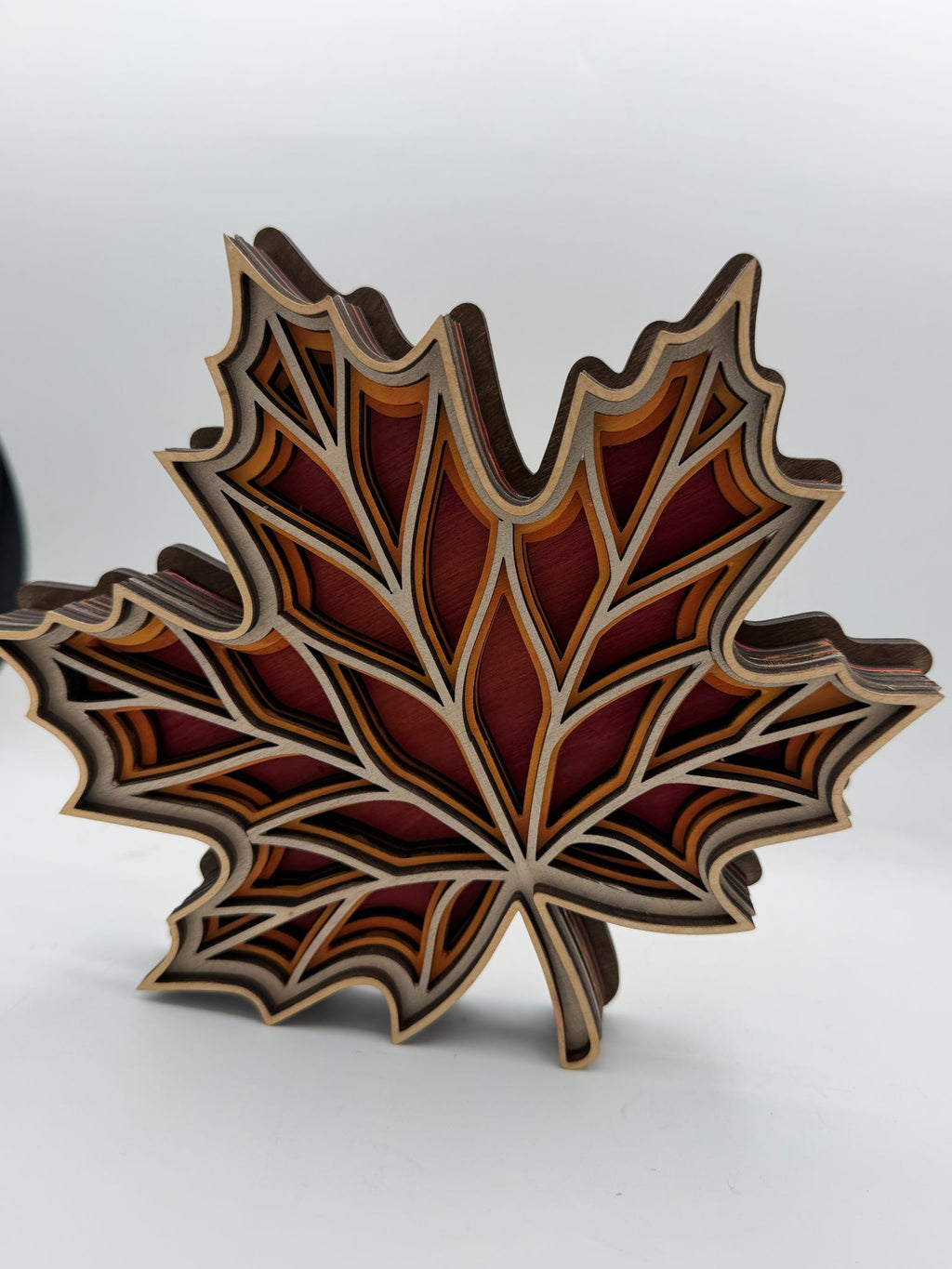 A 3D layered wooden maple leaf sculpture with intricate cutout details, showcasing warm tones of red, orange, brown, and beige wood grain against a neutral light gray background.