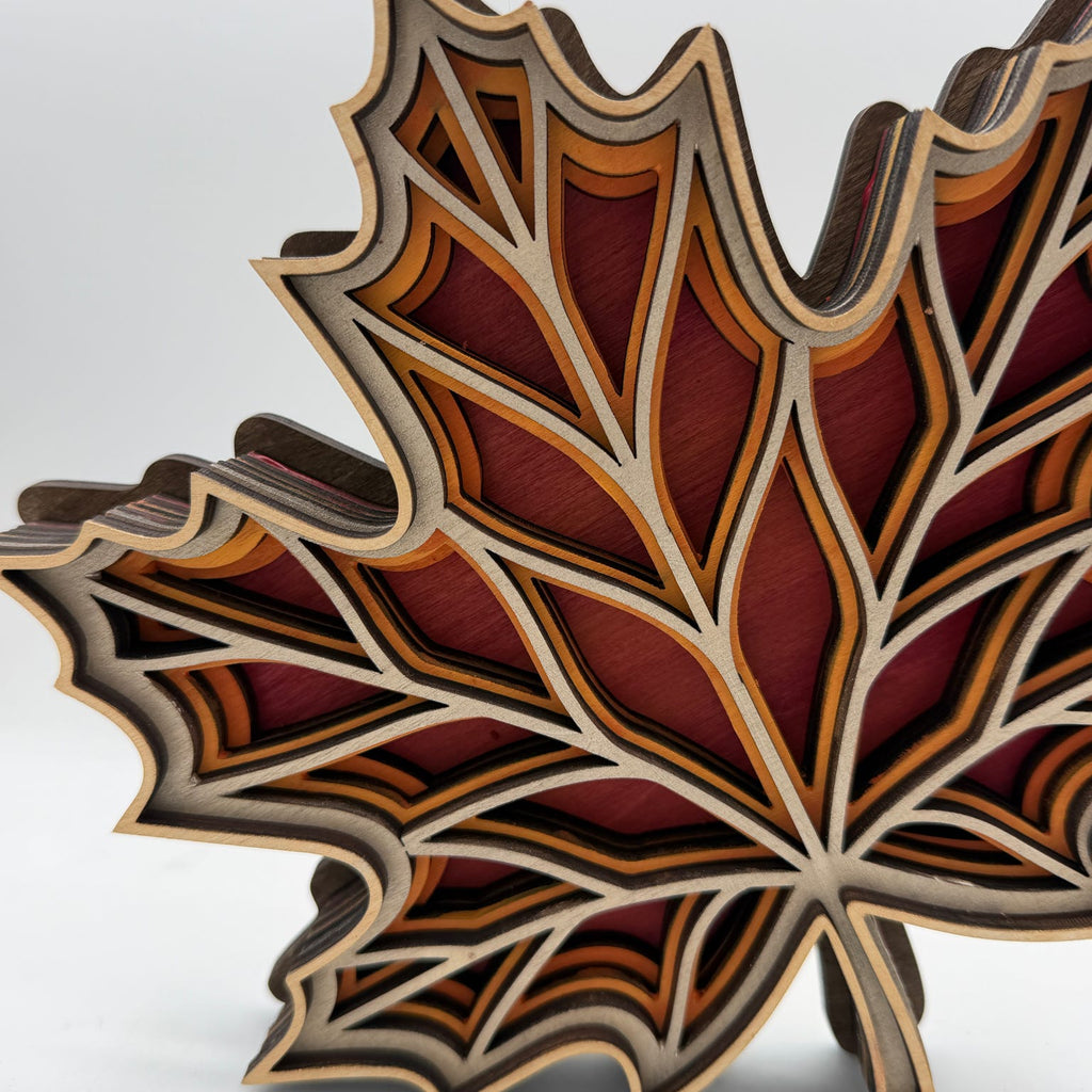 A 3D layered wooden leaf sculpture, likely shaped like a maple leaf, featuring intricate laser-cut layers in shades of red, brown, orange, and natural wood beige.