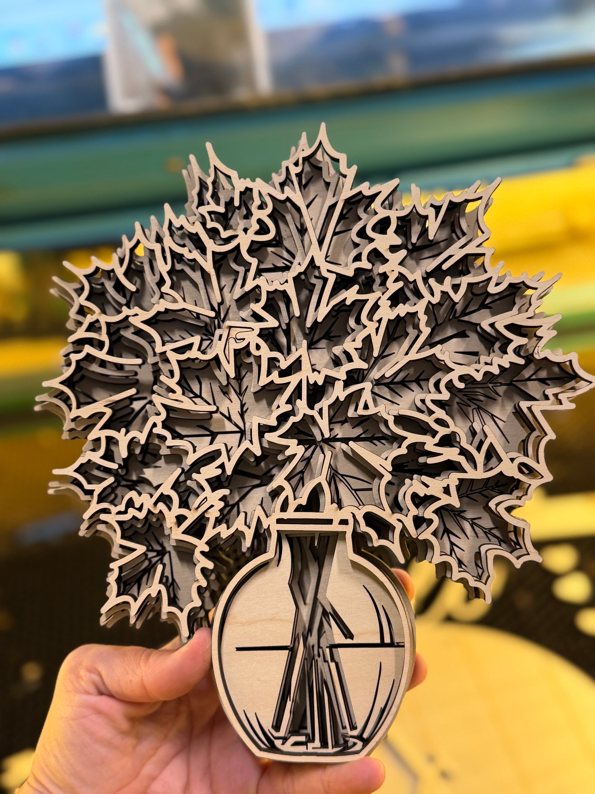A hand holds a laser-cut wooden art piece featuring a detailed maple leaf design in a vase, with a blurred outdoor background.