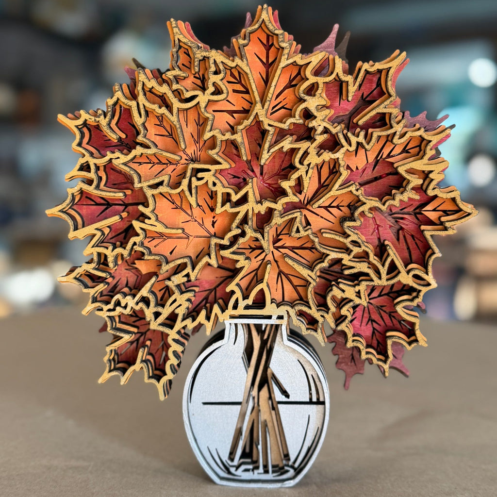 A 3D papercraft pop-up sculpture of autumn maple leaves in shades of orange, red, and brown, arranged in a white and black vase on a neutral surface with a blurred bokeh background.
