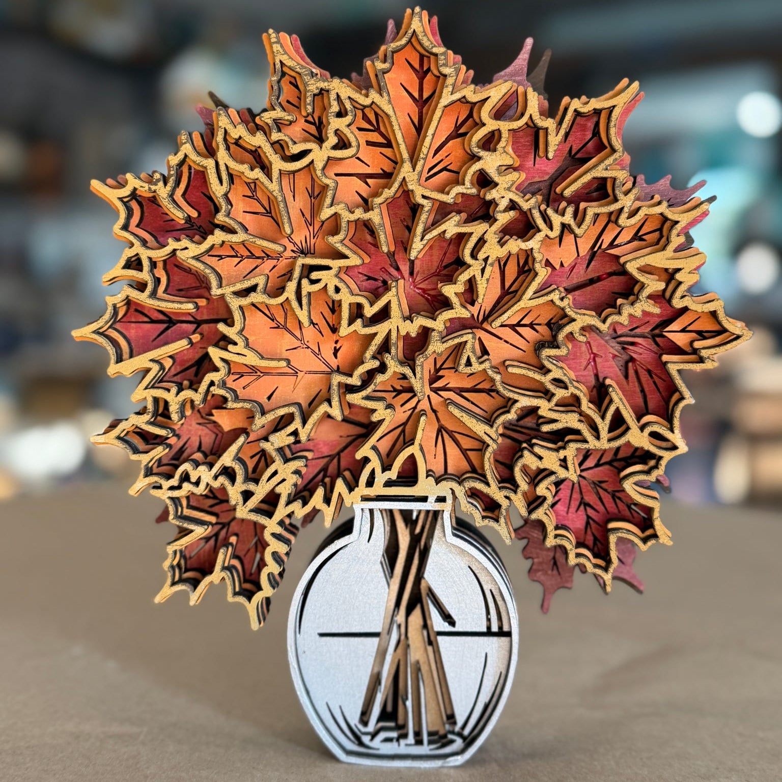 A 3D papercraft pop-up sculpture of autumn maple leaves in shades of orange, red, and brown, arranged in a white and black vase on a neutral surface with a blurred bokeh background.