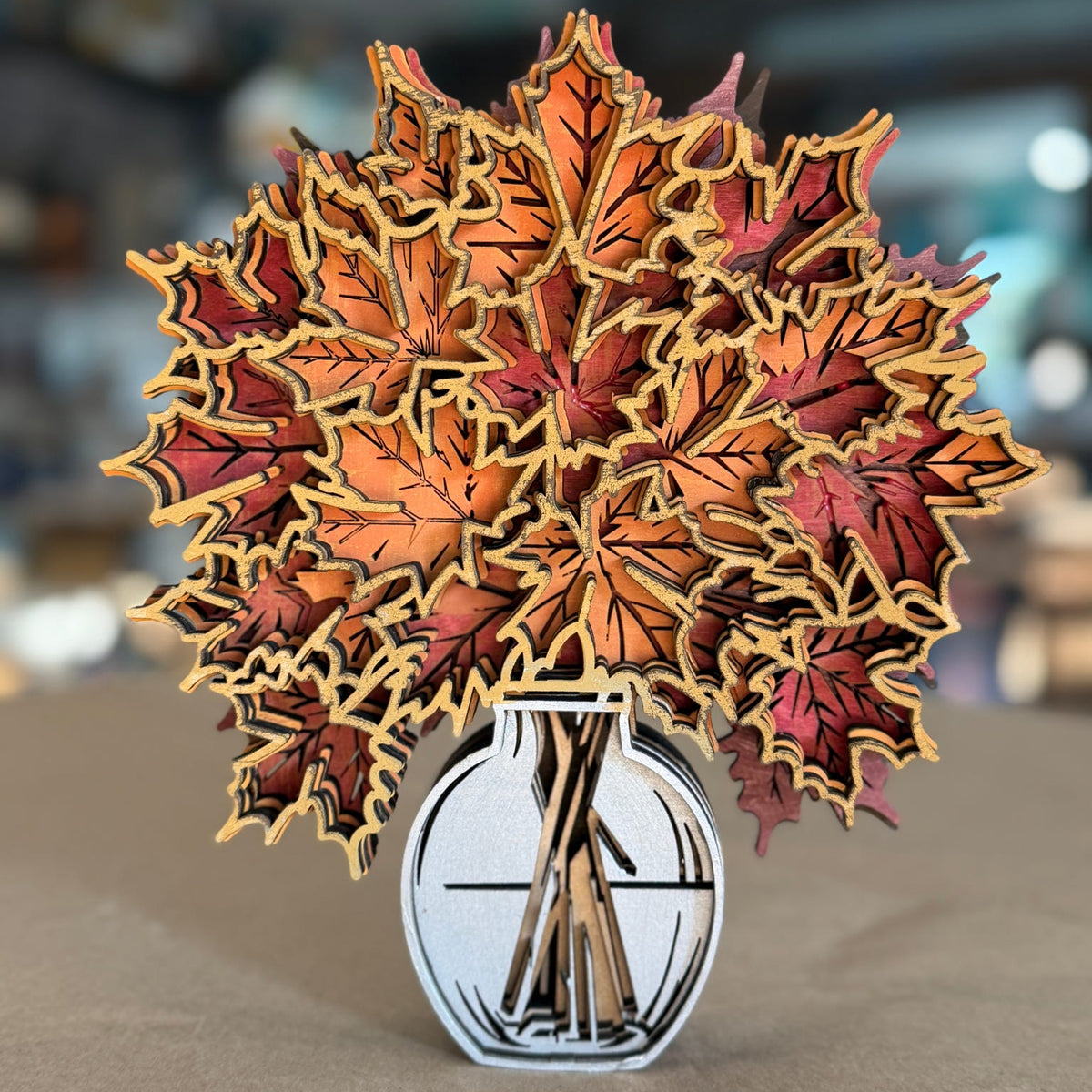 A 3D papercraft pop-up sculpture of autumn maple leaves in shades of orange, red, and brown, arranged in a white and black vase on a neutral surface with a blurred bokeh background.