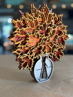 A 3D laser-cut wooden autumn leaf pop-up sculpture in a glass vase, displayed on a neutral surface with a blurred background.