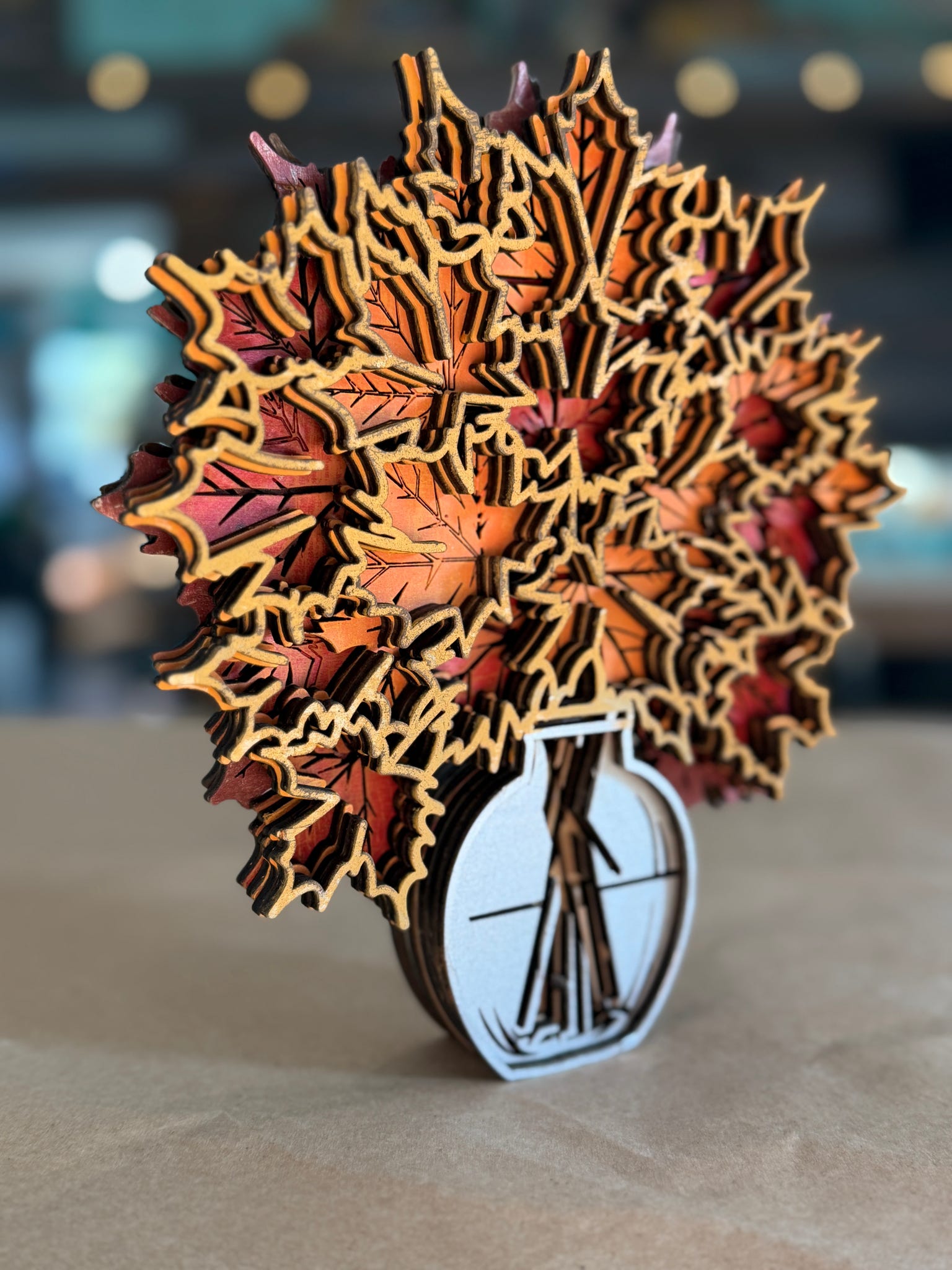 A 3D laser-cut wooden autumn leaf pop-up sculpture in a glass vase, displayed on a neutral surface with a blurred background.