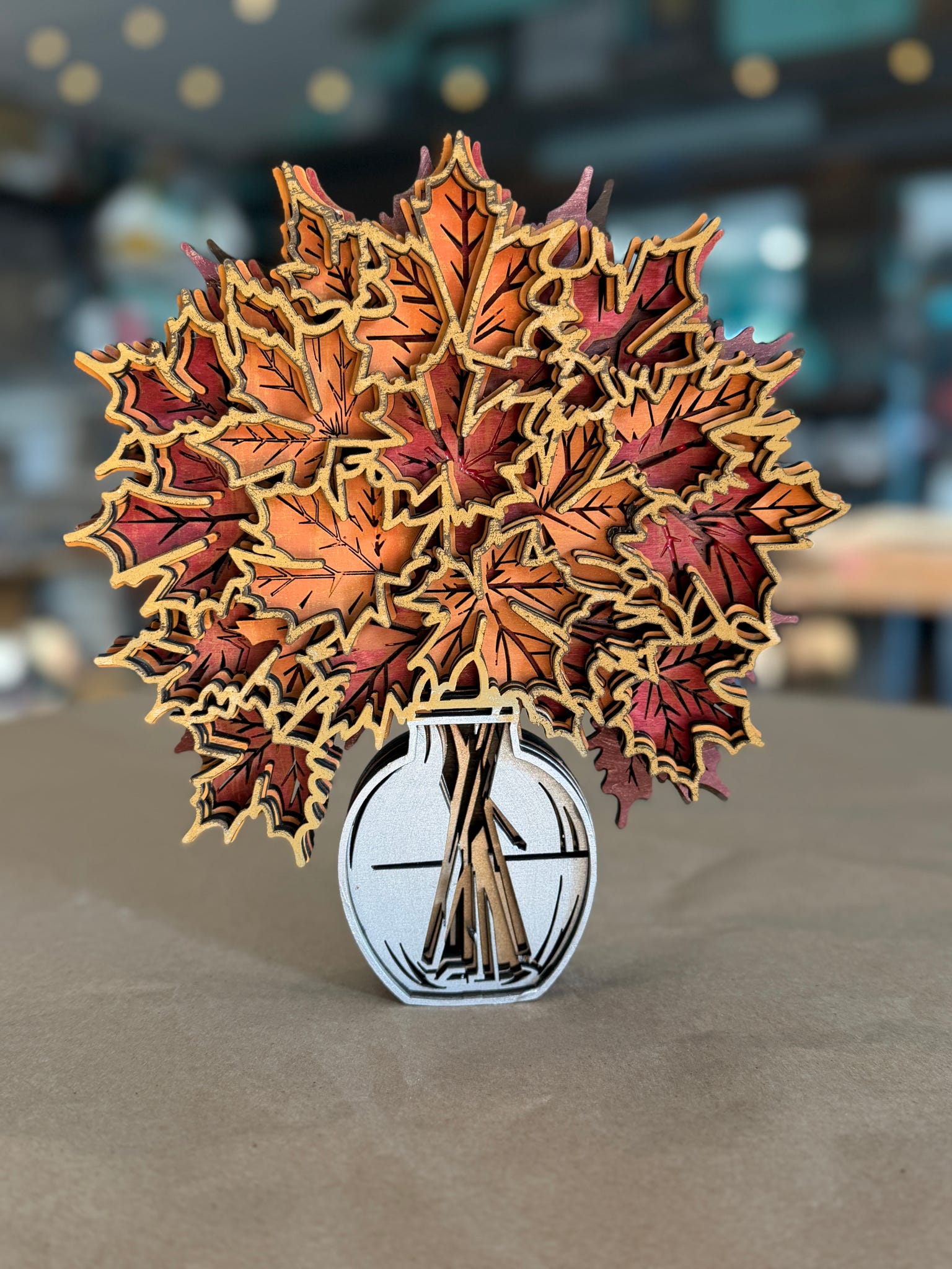 A decorative autumn leaf arrangement in a small white vase, photographed on a tabletop with a softly blurred indoor background.