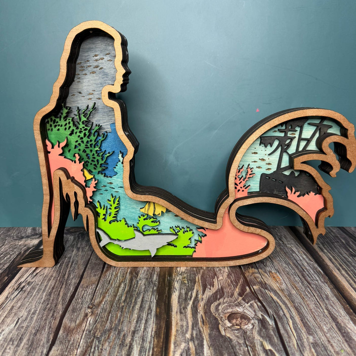 A decorative layered mermaid sign with a unique design, featuring a silhouette of a mermaid in multicolor layers, mounted on a wooden surface.