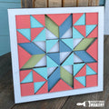 A layered quilt 3D sign with multiple geometric shaped pieces in blue, green, and pink colors assembled in a larger square frame.