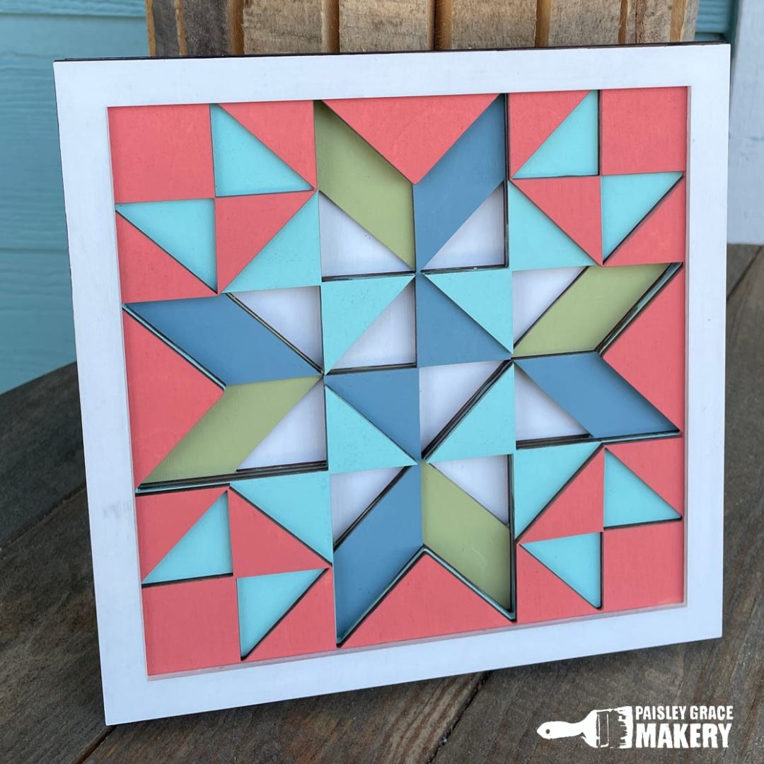 A layered quilt 3D sign with multiple geometric shaped pieces in blue, green, and pink colors assembled in a larger square frame.