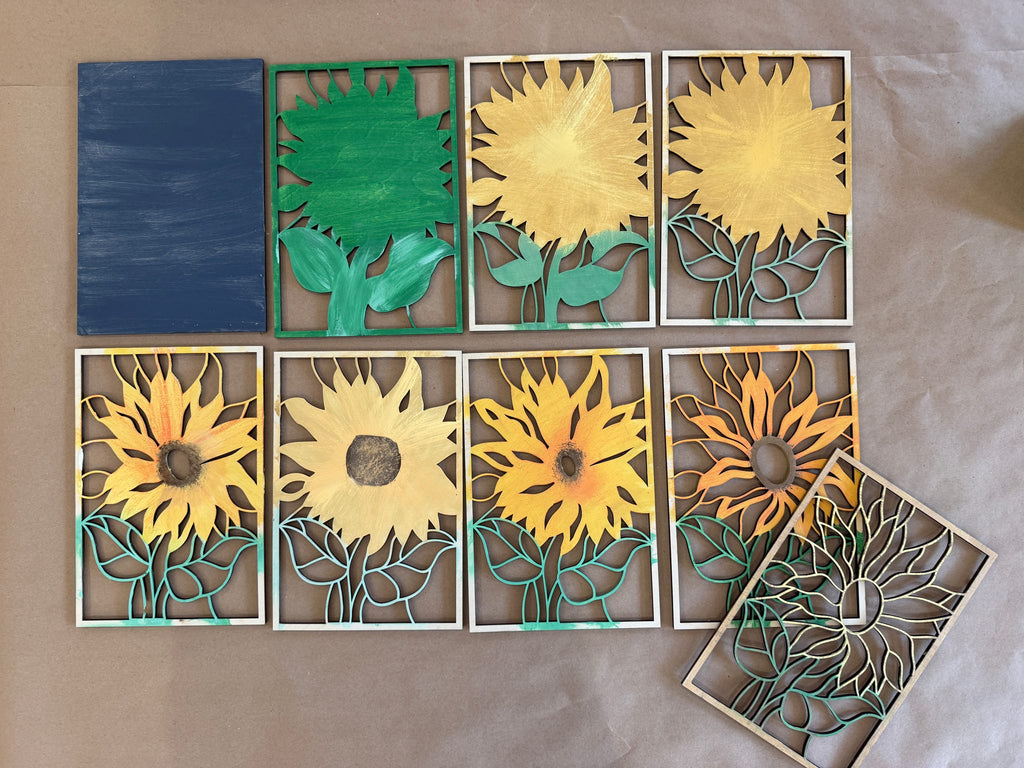 A set of eight handcrafted wooden sunflower wall art panels arranged on a beige surface. The panels feature various stages of a sunflower design, from a partially assembled wooden frame to fully painted yellow, green, and blue sunflower pieces.
