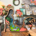 A wooden silhouette cutout sculpture of a mermaid standing on a wooden surface, with layered colorful ocean and coral scenes visible inside the cutout. The background is a softly blurred indoor shop or craft space.