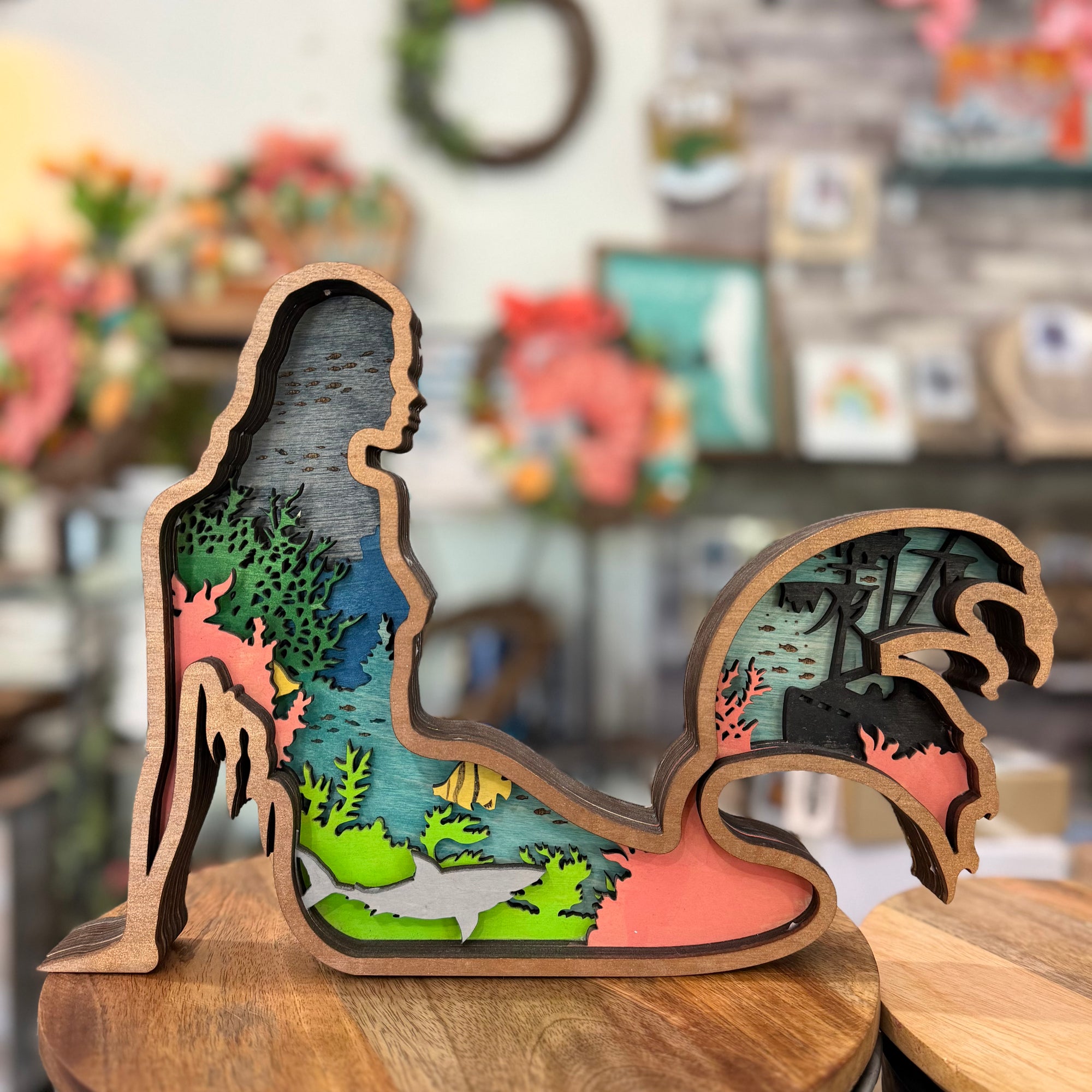 A wooden silhouette cutout sculpture of a mermaid standing on a wooden surface, with layered colorful ocean and coral scenes visible inside the cutout. The background is a softly blurred indoor shop or craft space.