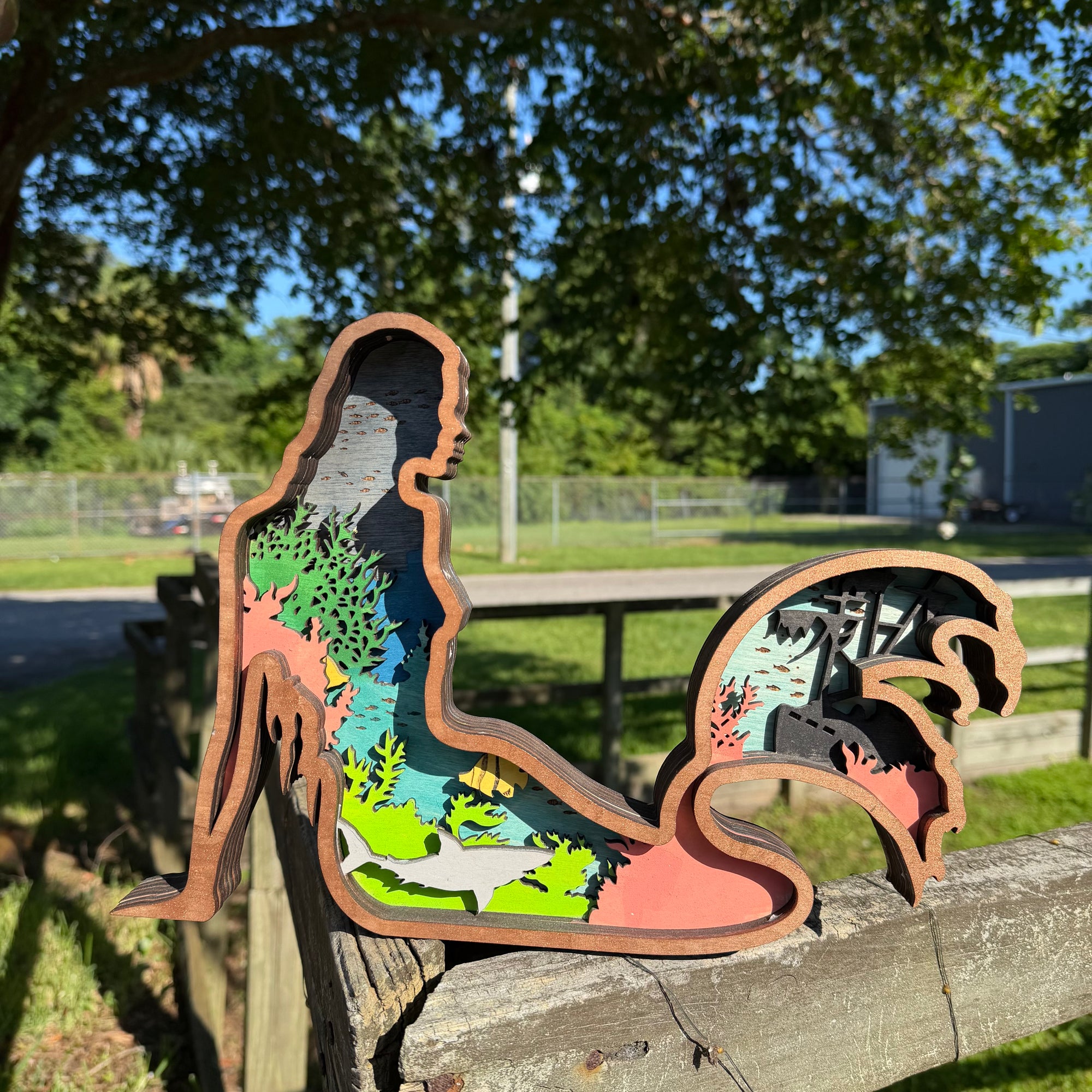 A wooden layered cutout sculpture of a seated figure with a wave-like tail, filled with colorful forest and landscape layers, photographed outdoors on a wooden fence with trees and a yard in the background.