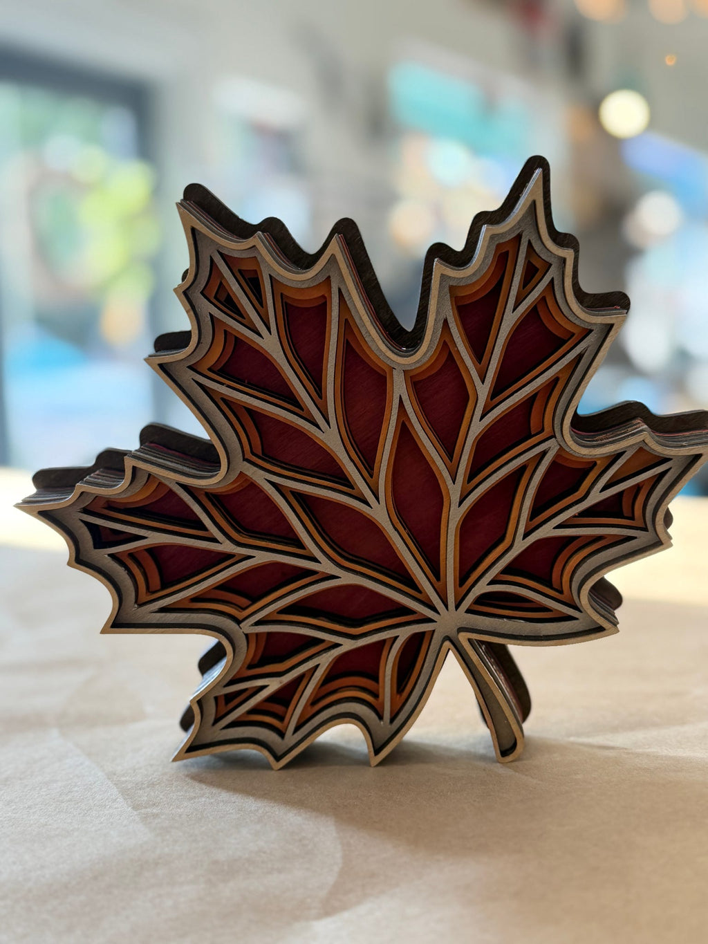 A decorative layered wooden maple leaf sculpture with red, brown, and beige tones, standing on a light surface with a blurred indoor background.