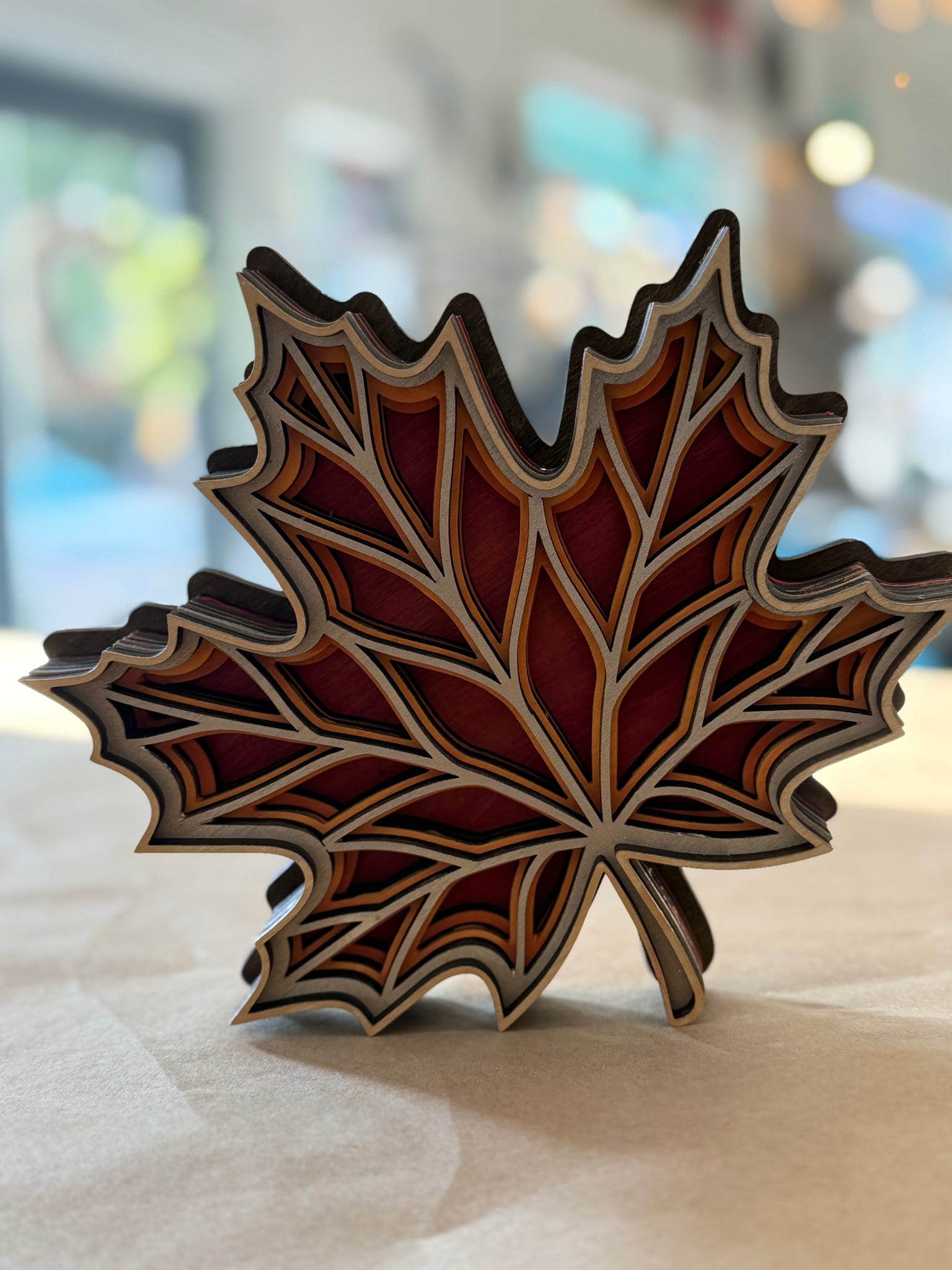A decorative layered wooden maple leaf sculpture with red, brown, and beige tones, standing on a light surface with a blurred indoor background.