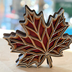 A decorative maple leaf-shaped object with layered wood or metal sections in red, brown, beige, and dark gray tones, photographed on a light surface with a blurred outdoor background.