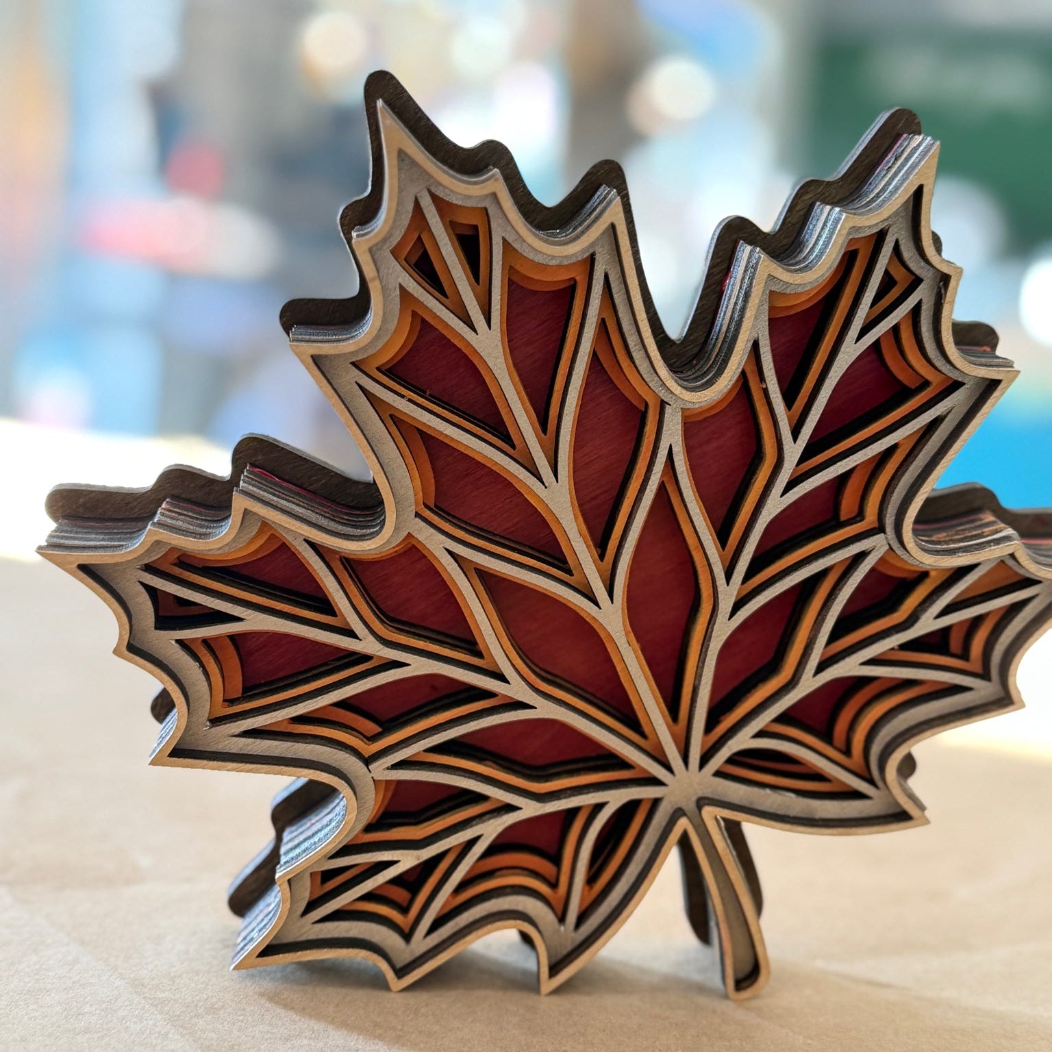 A decorative maple leaf-shaped object with layered wood or metal sections in red, brown, beige, and dark gray tones, photographed on a light surface with a blurred outdoor background.