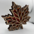 A 3D layered wooden maple leaf sculpture with a geometric design, showcasing natural wood grain and multiple colors of wood tones, set against a plain light gray background.