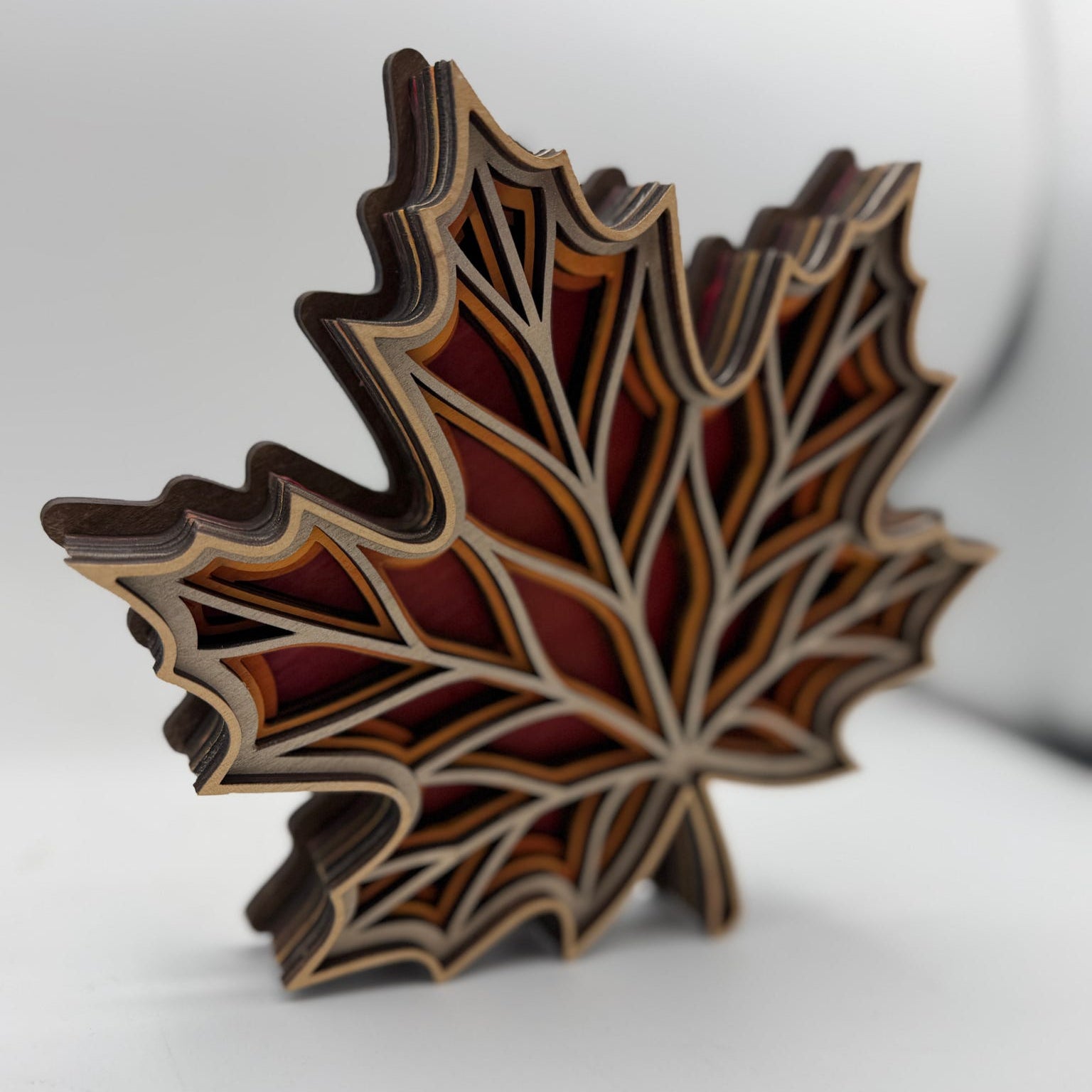 A 3D layered wooden maple leaf sculpture with a geometric design, showcasing natural wood grain and multiple colors of wood tones, set against a plain light gray background.