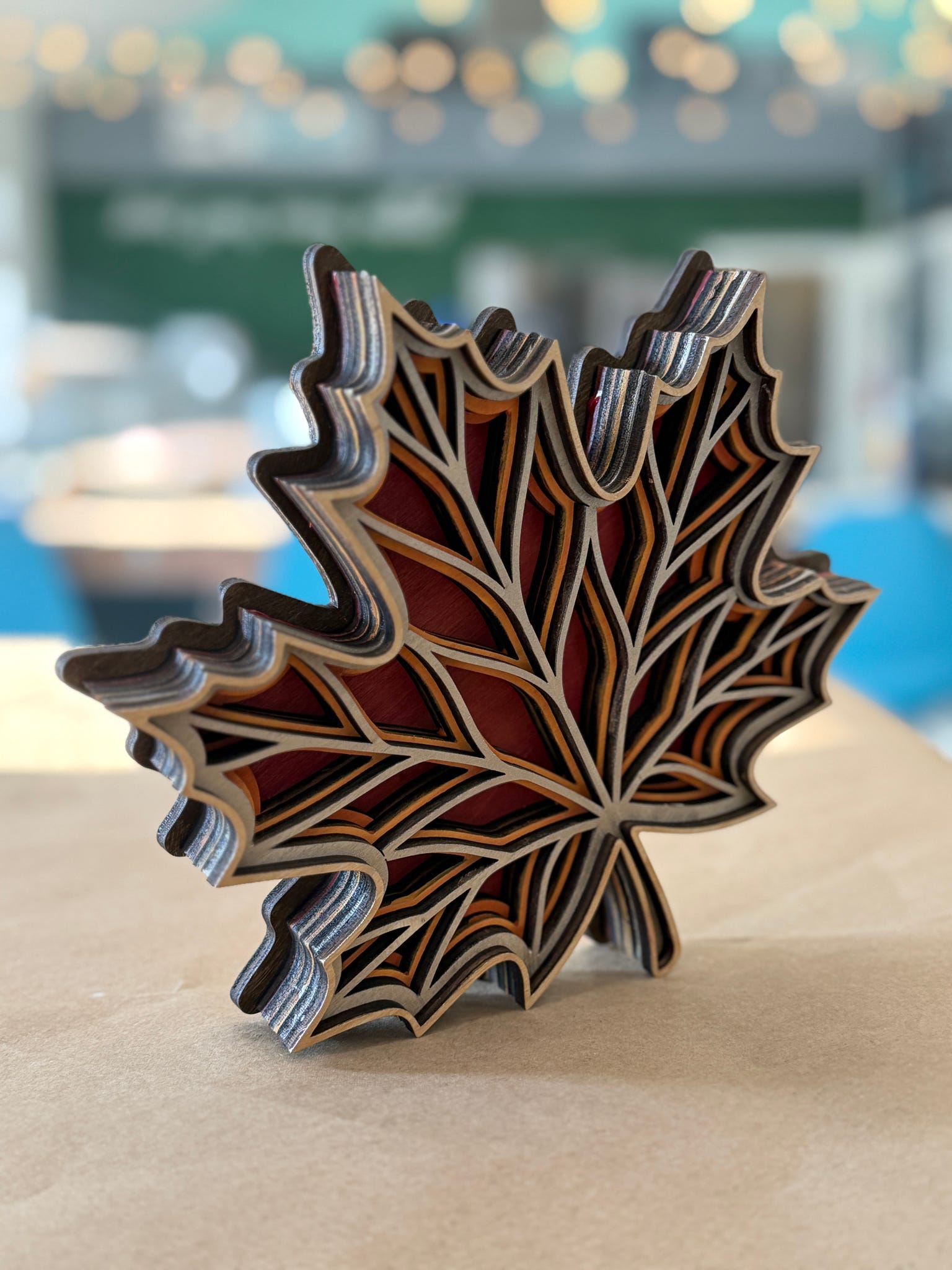 A 3D layered wooden leaf sculpture with a maple leaf design, featuring red, brown, and beige wood layers, standing on a light brown surface with a blurred background.