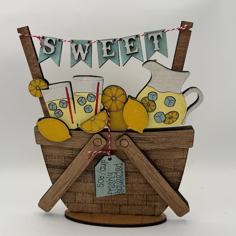 Decorative wooden basket with lemons, glasses, and a pitcher, featuring a 'Sweet' banner.