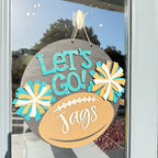 A round wooden hanging sign with a football design and the words “Let’s Go! Jags,” displayed on a glass door. The sign features turquoise and white pom-pom flower accents and is photographed with reflections of trees and sky in the window.