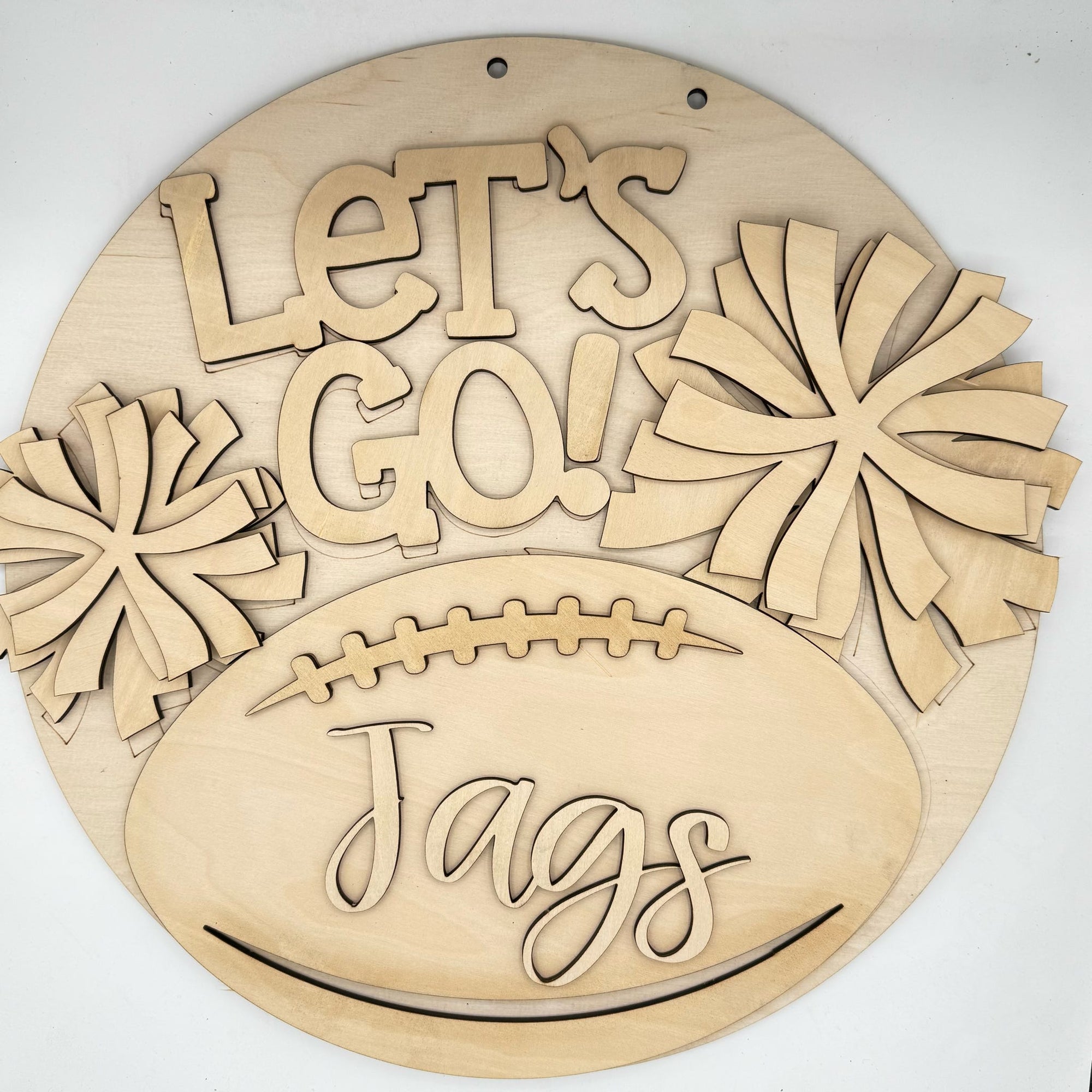 A round wooden wall sign with laser-cut text reading 'Let's Go! Jags' and pom-pom shapes, displayed on a plain white background.