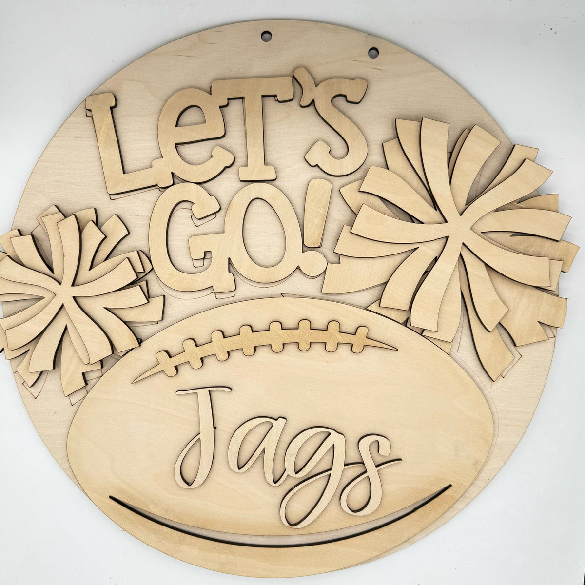 A round wooden wall sign with laser-cut text reading 'Let's Go! Jags' and pom-pom shapes, displayed on a plain white background.