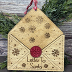 Wooden ornament shaped like a letter with snowflakes and a red wax seal, hanging on a wooden surface with greenery.