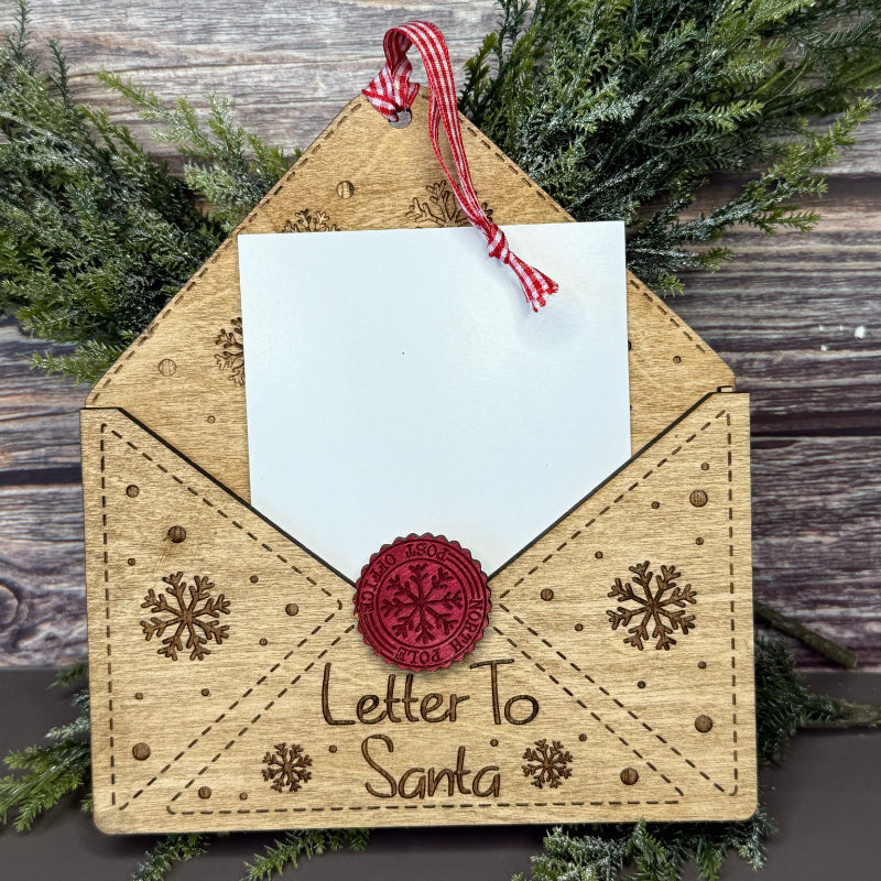 Wooden envelope ornament with 'Letter To Santa' text and snowflake designs, tied with a red and white string against a wooden background.