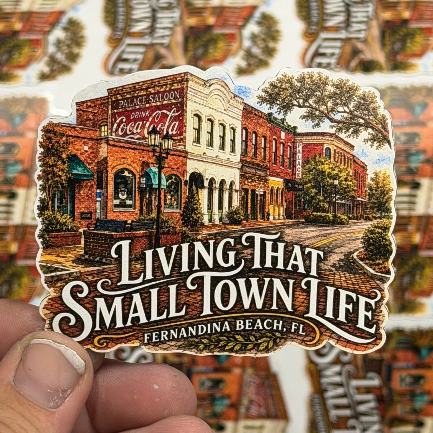 Sticker of a small town scene with 'Living That Small Town Life' text, held over a sheet of similar stickers.