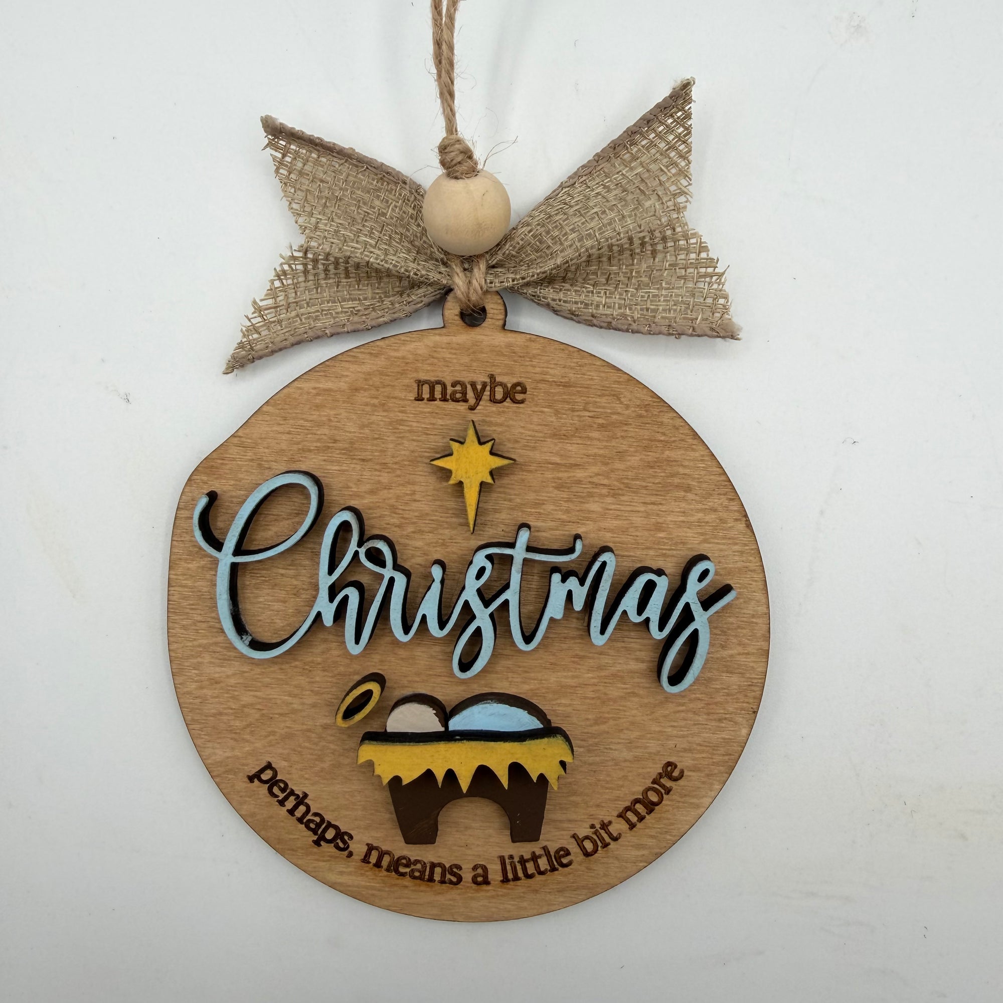 A round wooden Christmas ornament hanging from a burlap bow and wooden bead. The ornament is engraved with the words 'maybe Christmas perhaps means a little bit more' and features a small gold star and nativity scene.