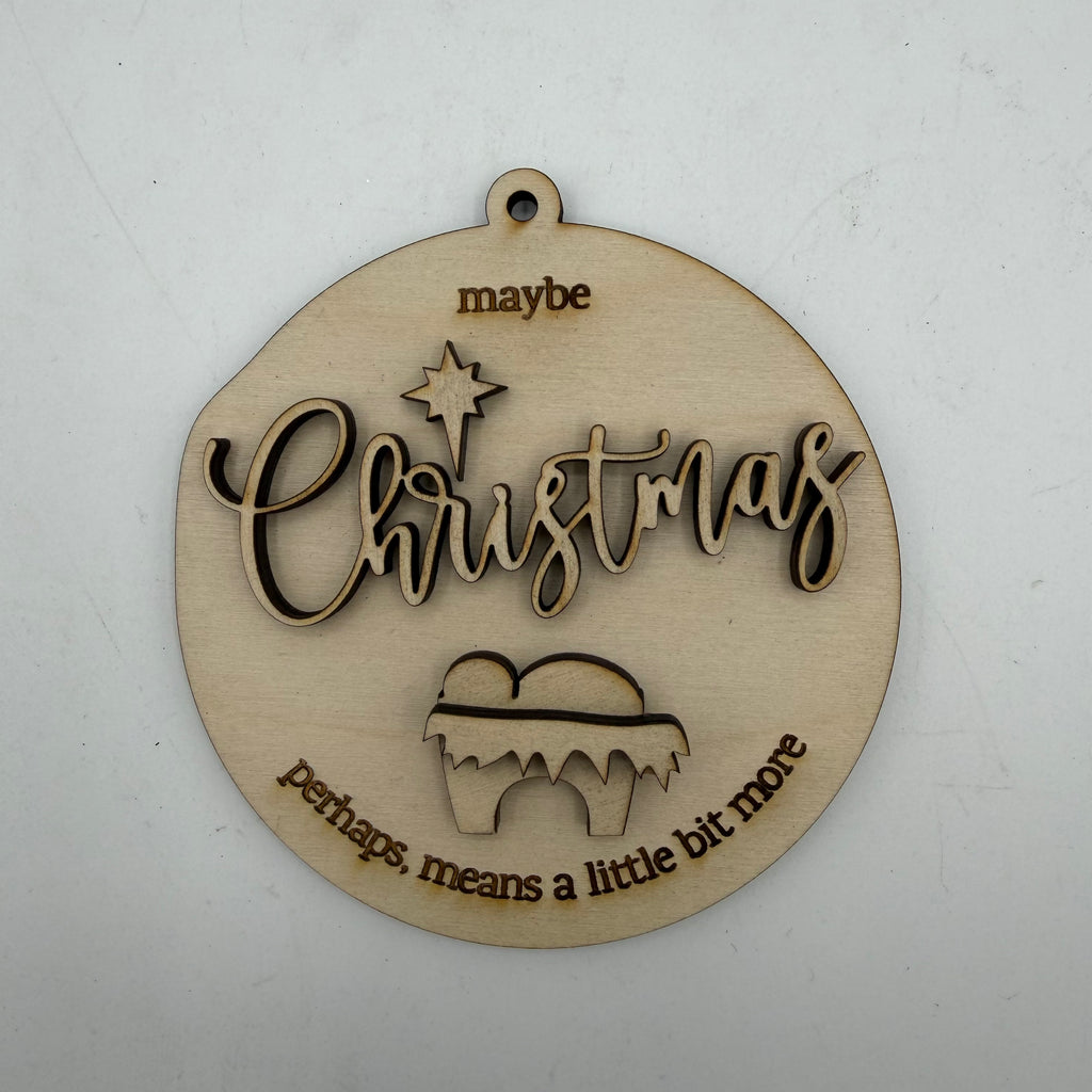 A round, laser-cut wooden Christmas ornament with a hanging loop at the top. The ornament features the word 'Christmas' in cursive with a star above it, and the phrases 'maybe' at the top and 'perhaps, means a little bit more' at the bottom, all in a light natural wood finish on a white background.