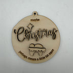 A round, laser-cut wooden Christmas ornament with a hanging loop at the top. The ornament features the word 'Christmas' in cursive with a star above it, and the phrases 'maybe' at the top and 'perhaps, means a little bit more' at the bottom, all in a light natural wood finish on a white background.