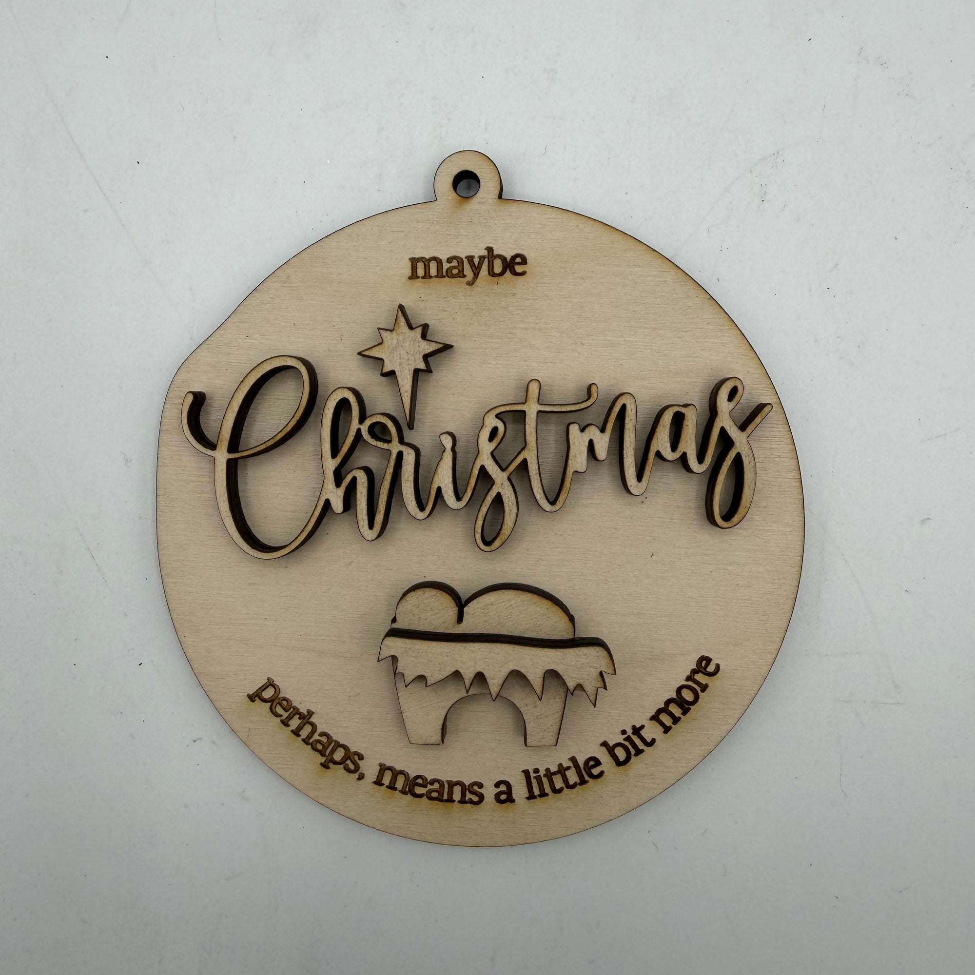 A round, laser-cut wooden Christmas ornament with a hanging loop at the top. The ornament features the word 'Christmas' in cursive with a star above it, and the phrases 'maybe' at the top and 'perhaps, means a little bit more' at the bottom, all in a light natural wood finish on a white background.