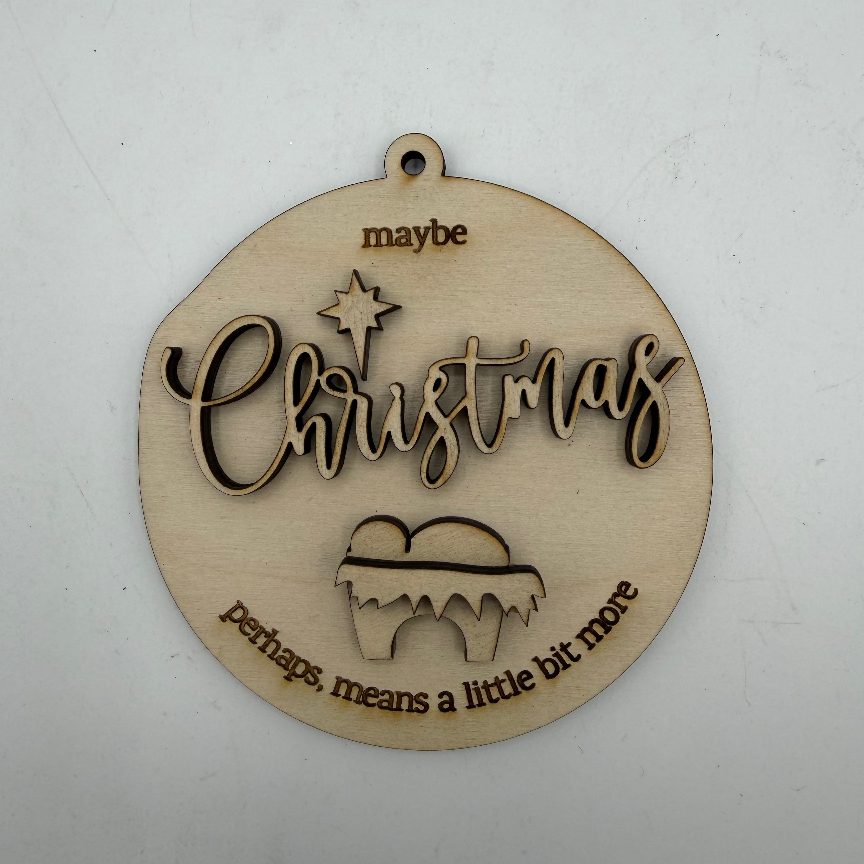 A round, laser-cut wooden Christmas ornament with a hanging loop at the top. The ornament features the word 'Christmas' in cursive with a star above it, and the phrases 'maybe' at the top and 'perhaps, means a little bit more' at the bottom, all in a light natural wood finish on a white background.