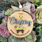 A round wooden Christmas ornament with a burlap bow and hanging loop, featuring the words “maybe Christmas perhaps means a little bit more” and a small nativity scene, displayed among purple and green succulents.