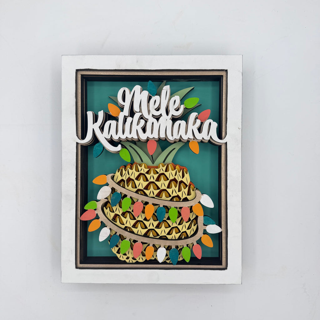 A square framed wall art piece featuring a stylized pineapple with the words “Mele Kalikok’a” in white script across the top, set against a teal background with multicolored string lights and a white frame.