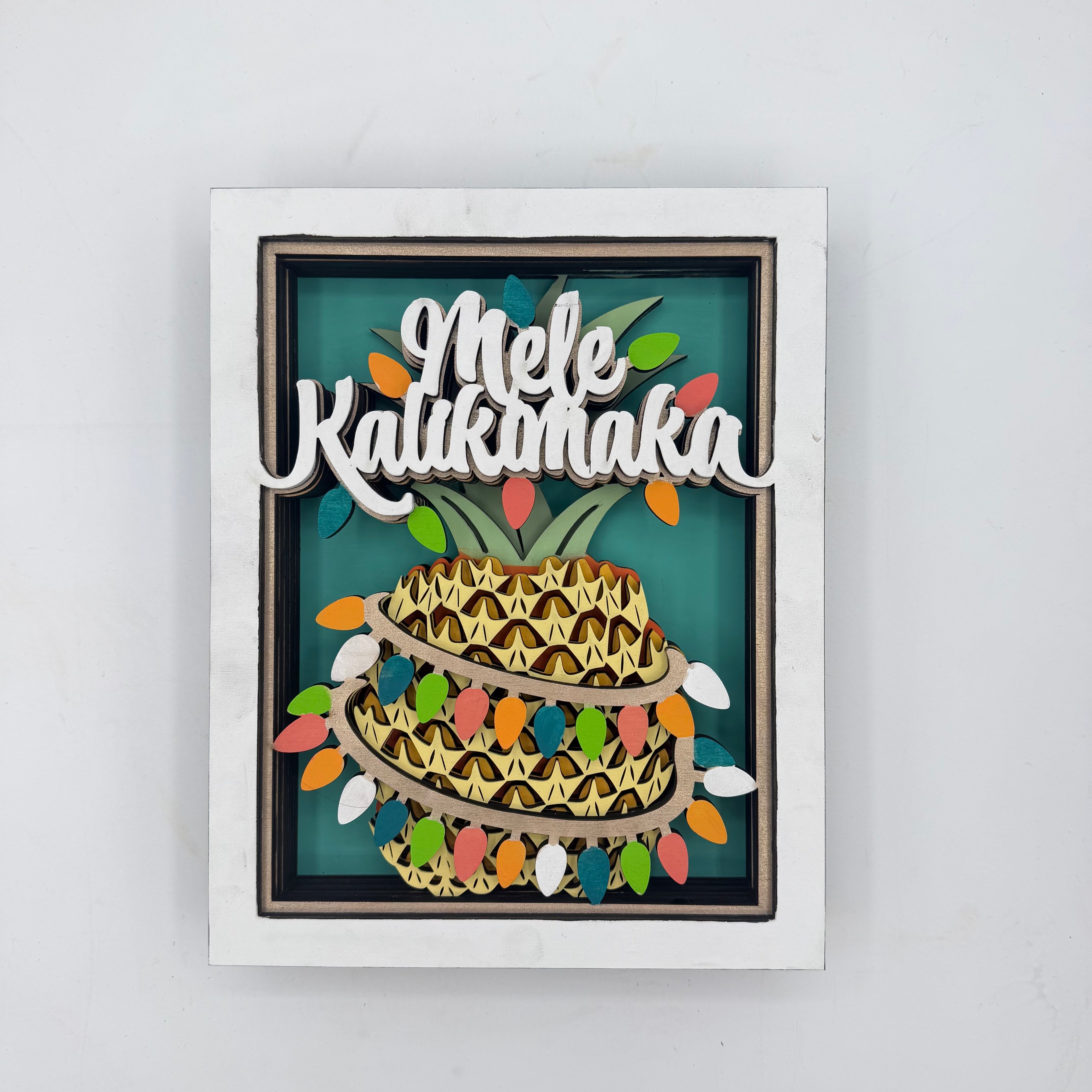A square framed wall art piece featuring a stylized pineapple with the words “Mele Kalikok’a” in white script across the top, set against a teal background with multicolored string lights and a white frame.